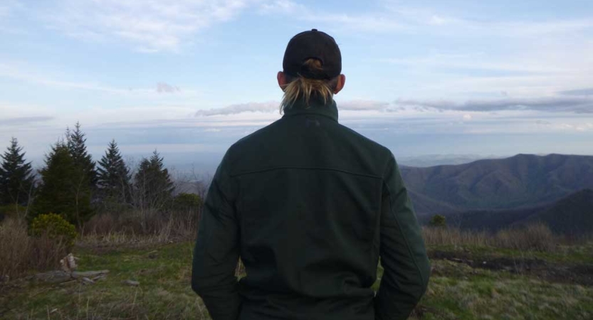 A person looks away from the camera, out toward a mountainous landscape and evergreen trees